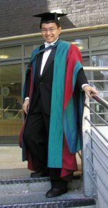 Man in academic robes (teal, with red and teal sleeves), a black suit and a mortarboard. 